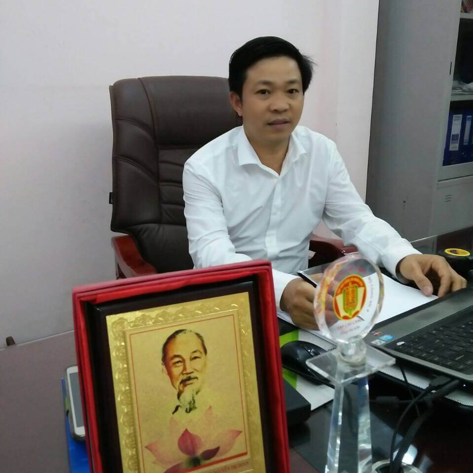 Người đàn ông mặc sơ mi trắng ngồi tại bàn làm việc, bên cạnh là kỷ niệm chương và ảnh Chủ tịch Hồ Chí Minh